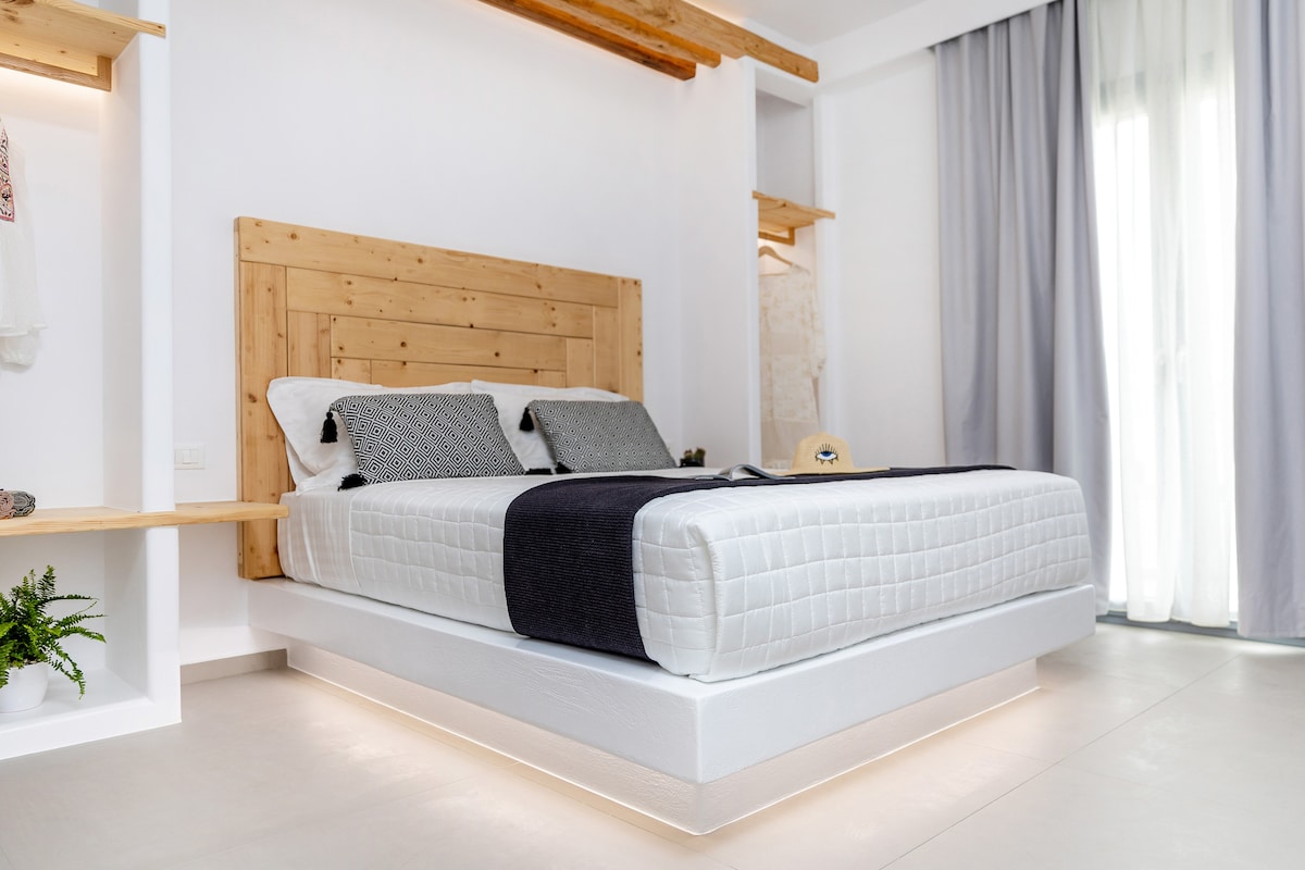 A spacious bed is set against a wooden headboard, featuring neutral linens and accented by two decorative pillows. Natural light enters through sheer curtains, illuminating the light-colored floor and minimalist decor, which includes a shelf and hanging space.