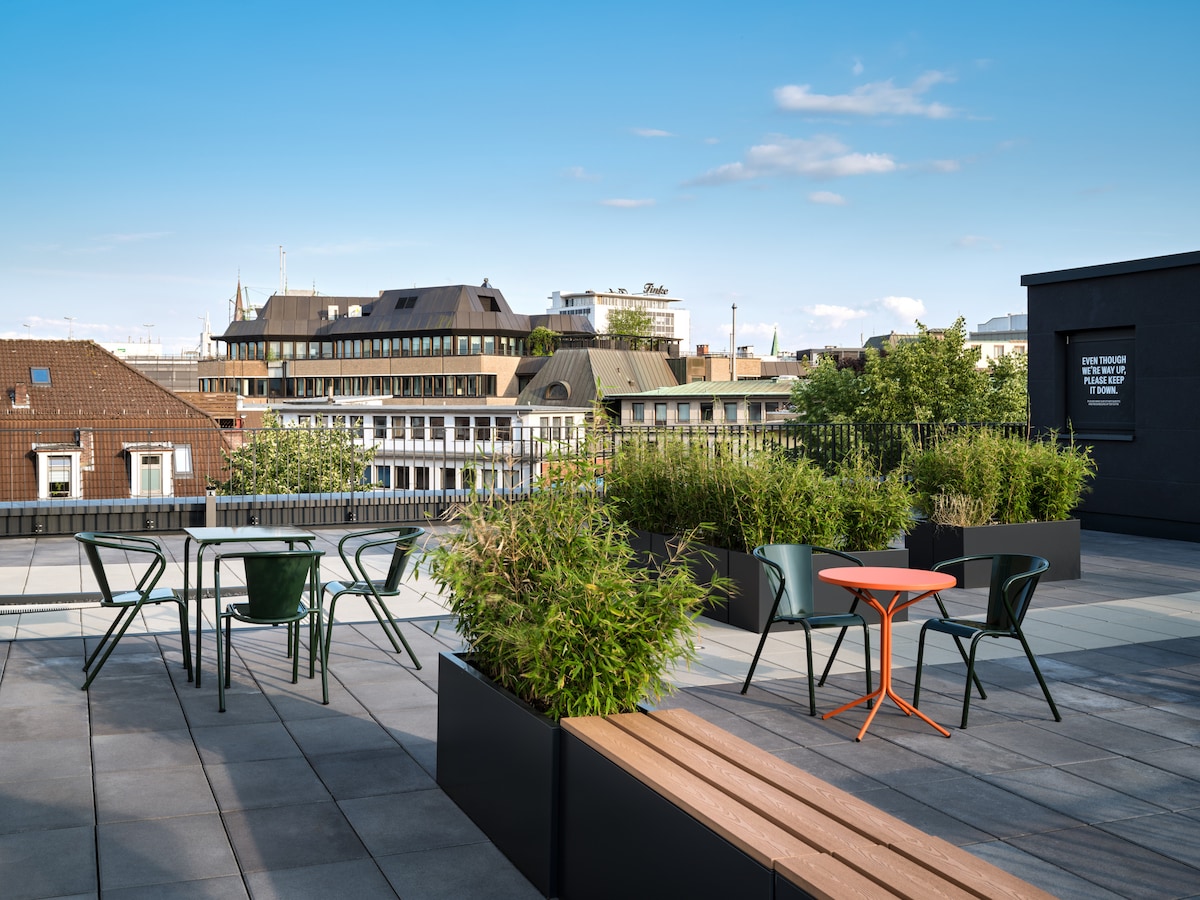 A rooftop terrace is displayed, featuring modern outdoor furniture. Green planters with lush greenery surround a round green table and black chairs, along with an orange side table. An expansive city skyline remains visible in the background under a clear blue sky.
