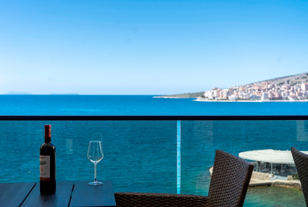 A serene view of the sea is framed by a glass railing, with a bottle of red wine and a glass resting on a wooden table. The vibrant blue water extends into the distance, while a coastal town is visible along the shore.