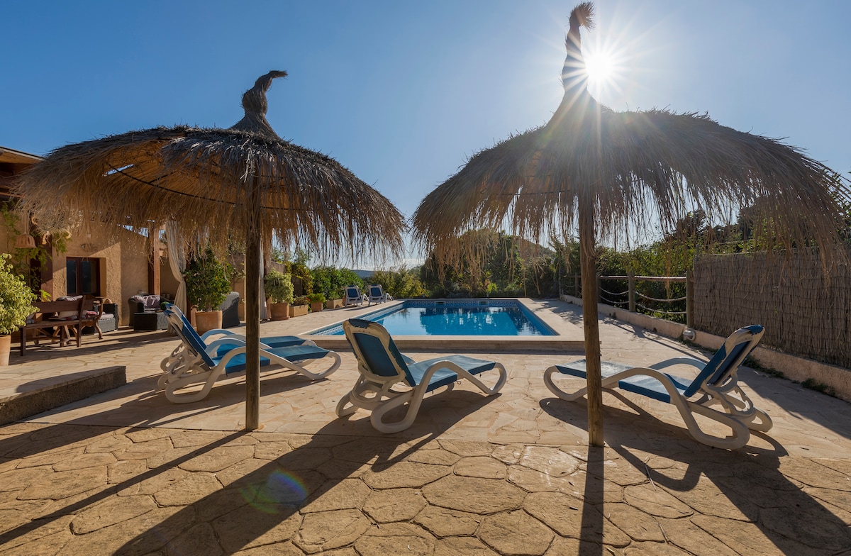 A sunny pool area is shaded by two thatched umbrellas, providing a relaxing spot near the water. Lounge chairs are placed on the stone patio, with vibrant green foliage visible in the background. The pool reflects sunlight under a clear blue sky.