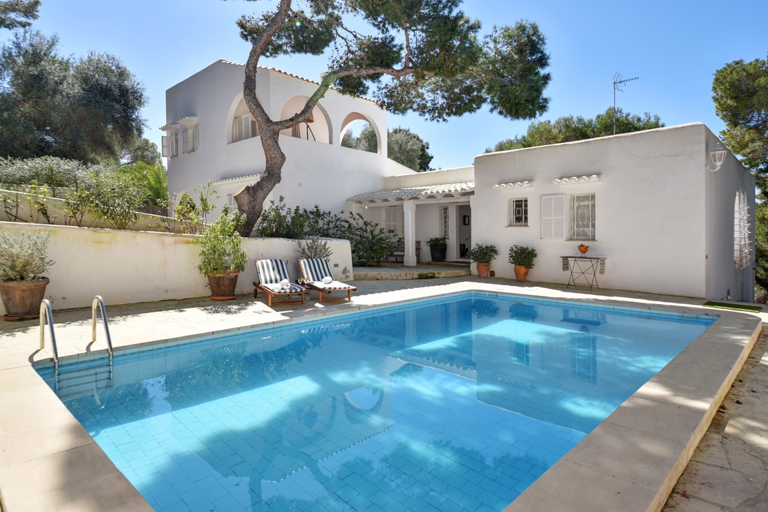 A clear blue swimming pool is framed by a white-walled villa and lush greenery. Two deck chairs are positioned beside the pool, inviting relaxation. The building features arched windows and a shaded terrace, creating a welcoming outdoor space.