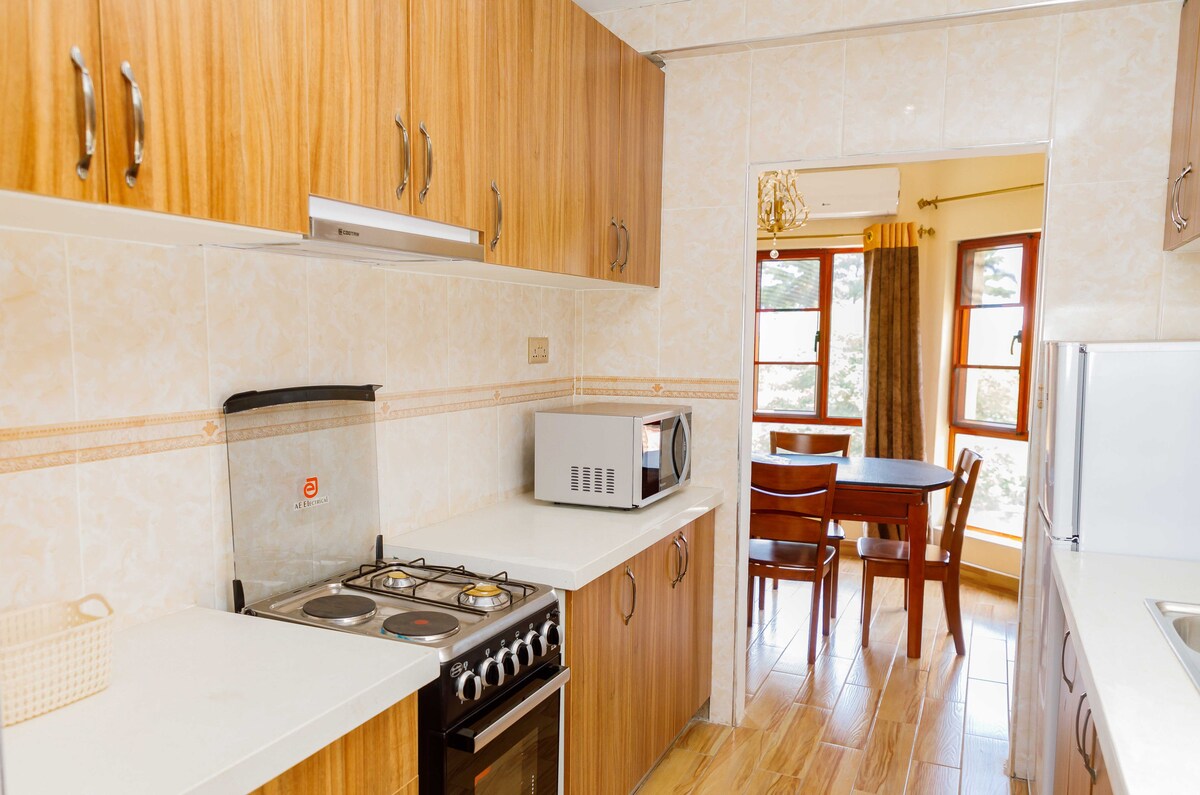 A well-equipped kitchen features wooden cabinets and a light countertop. A gas stove and microwave are visible, alongside a small basket. Adjacent to the kitchen, a dining table with four chairs is situated near large windows, allowing natural light to brighten the space.