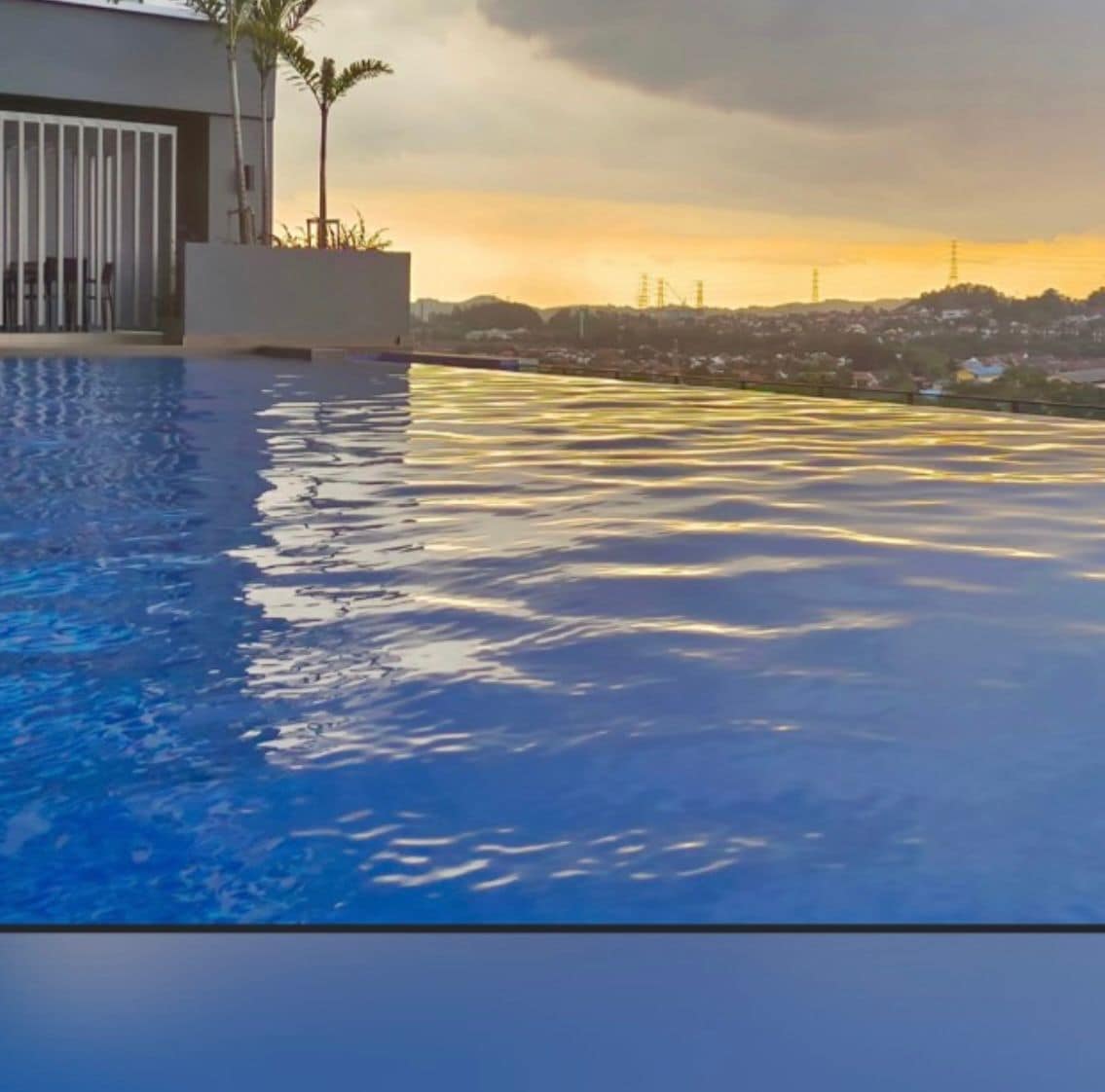 An infinity pool reflects the colorful hues of sunset, with gentle ripples across its surface. In the background, lush hills and a distant skyline are visible, while a small seating area is subtly positioned nearby under a few slender palm trees.