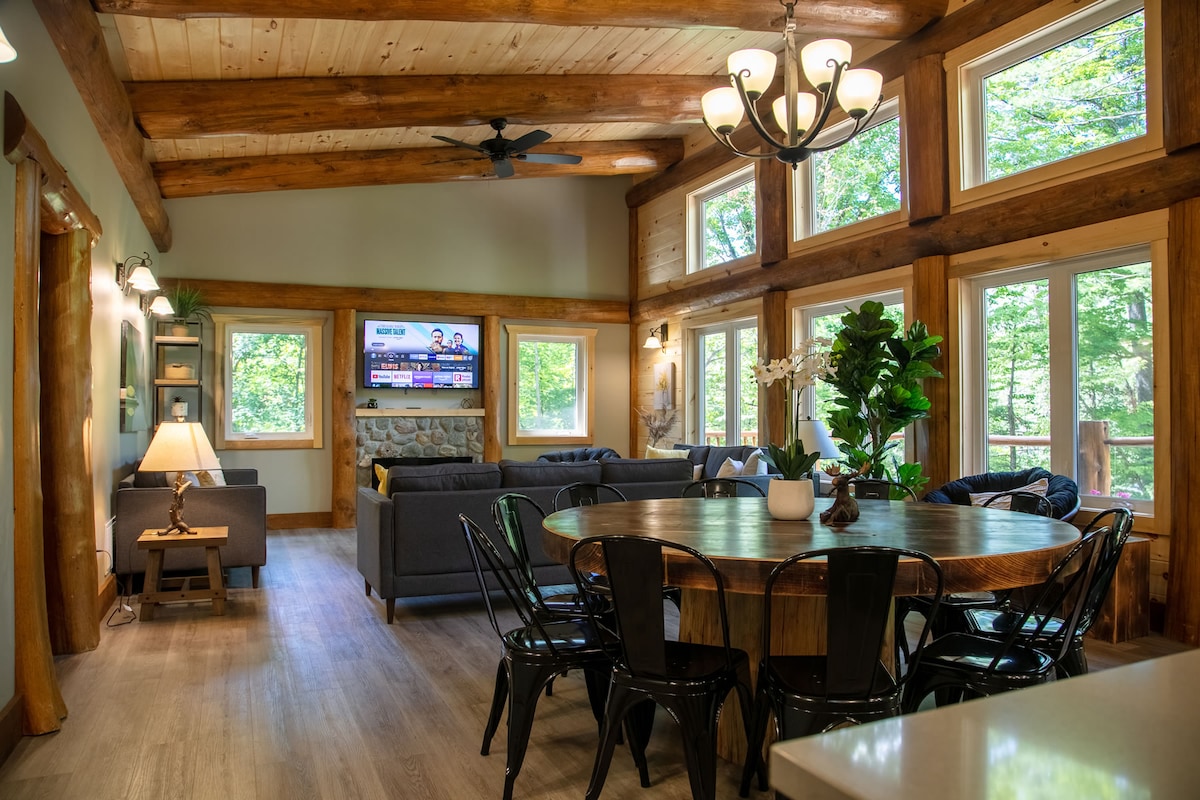 The spacious great room showcases high ceilings with exposed wood beams, large windows providing ample natural light, and a cozy seating area facing a television. A round wooden dining table is surrounded by black metal chairs, creating an inviting space for gatherings.