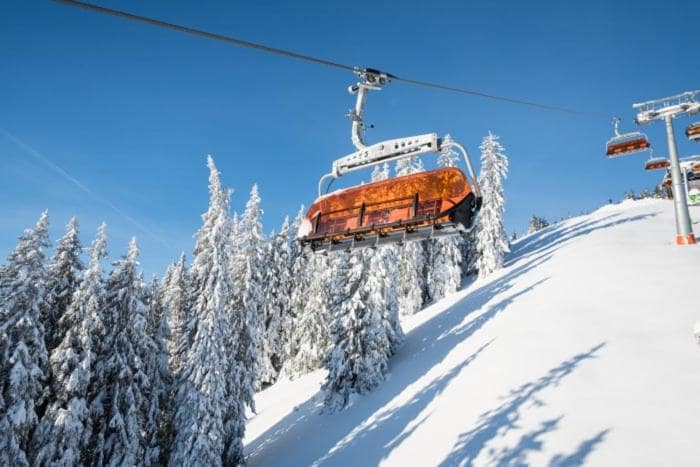 A ski lift is seen ascending a snow-covered slope surrounded by tall evergreen trees. The clear blue sky above contrasts with the white snow, creating a serene winter landscape. The lift cabins are visible in orange, offering a striking appearance against the natural backdrop.