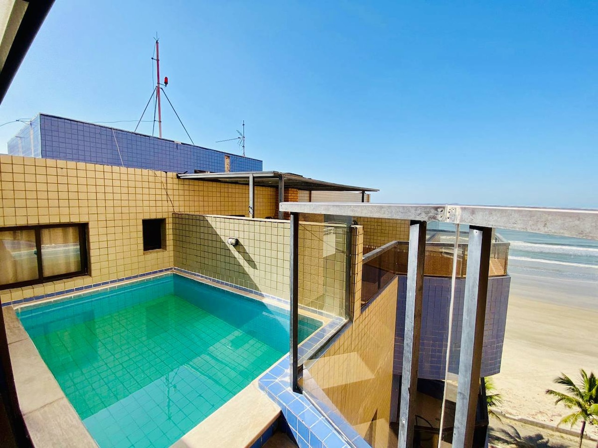 A private pool is visible on the balcony of a duplex, surrounded by glass railing. The clear blue water reflects the sky, providing a serene atmosphere. The ocean can be seen in the background, with the sandy beach just beyond.