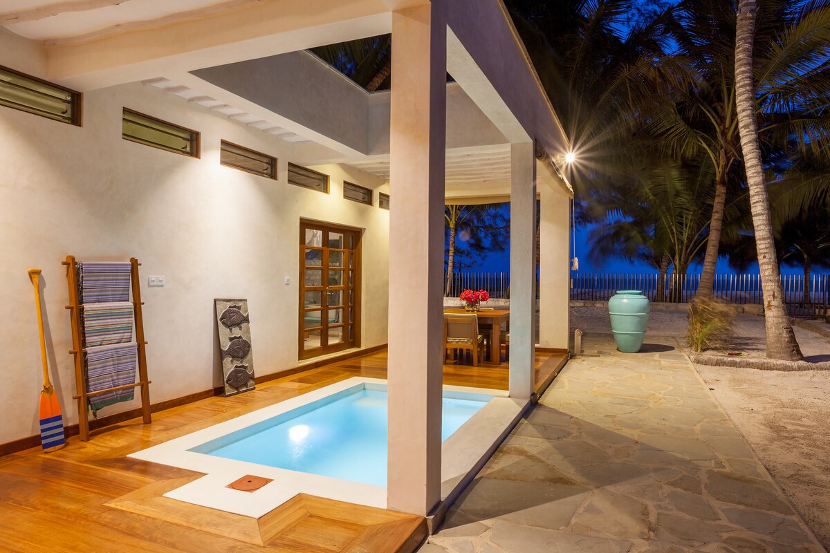 An open outdoor area features a private plunge pool, surrounded by natural stone flooring. Nearby, a dining table is set with bright flowers, complemented by coastal decor. Soft lighting creates a calming ambiance, with palm trees visible in the background under a twilight sky.