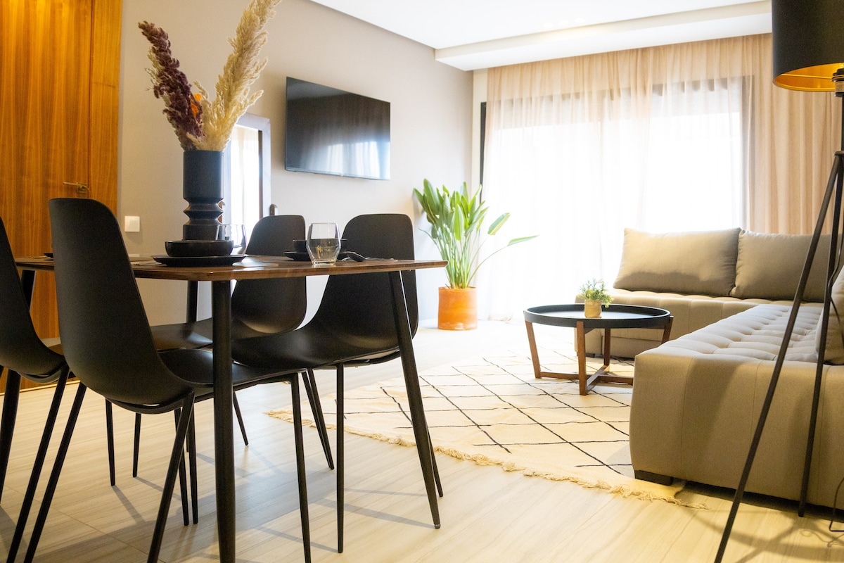 A modern living area is displayed with a wooden dining table surrounded by black chairs. A sofa is positioned nearby, complemented by decorative plants and a coffee table. Soft natural light filters through large windows, enhancing a relaxed atmosphere.