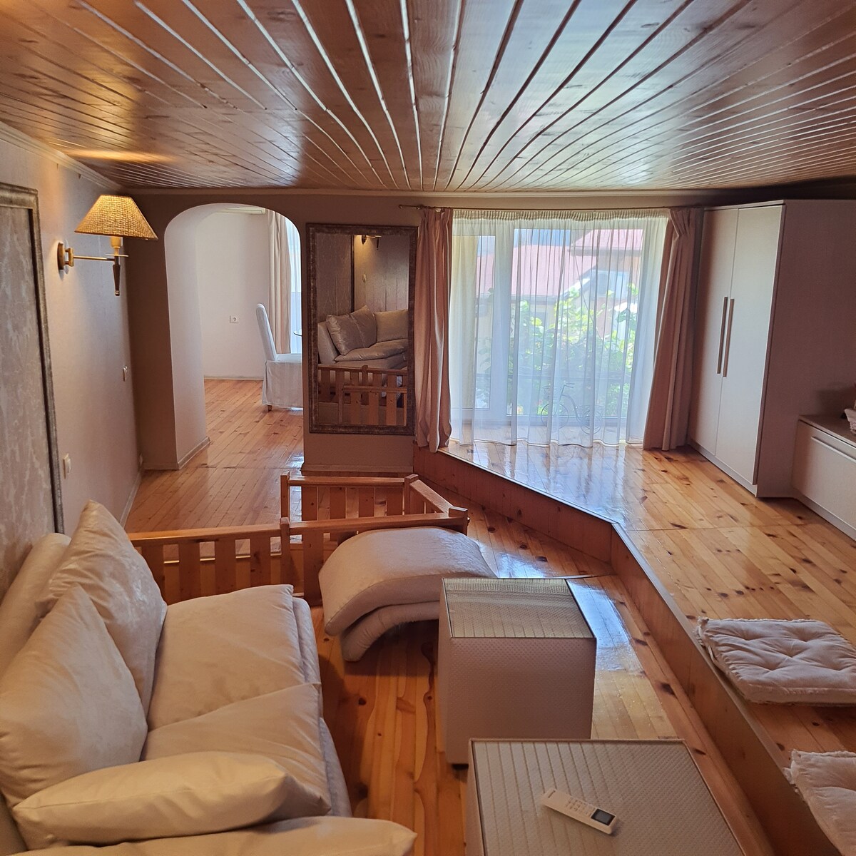 An inviting living area features light wooden flooring and a beamed ceiling. Soft seating options and textured pillows create a relaxed environment. Natural light filters through large windows, illuminating the space. A mirror enhances the sense of openness and reflects the greenery outside.