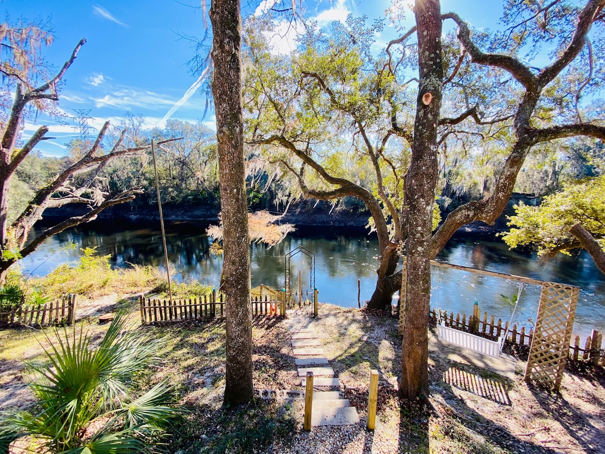 A view of the tranquil Withlacoochee River is framed by mature trees and greenery. Steps lead down to the water's edge, where natural beauty surrounds the inviting shoreline. Sunlight reflects off the calm water, creating a peaceful outdoor setting.