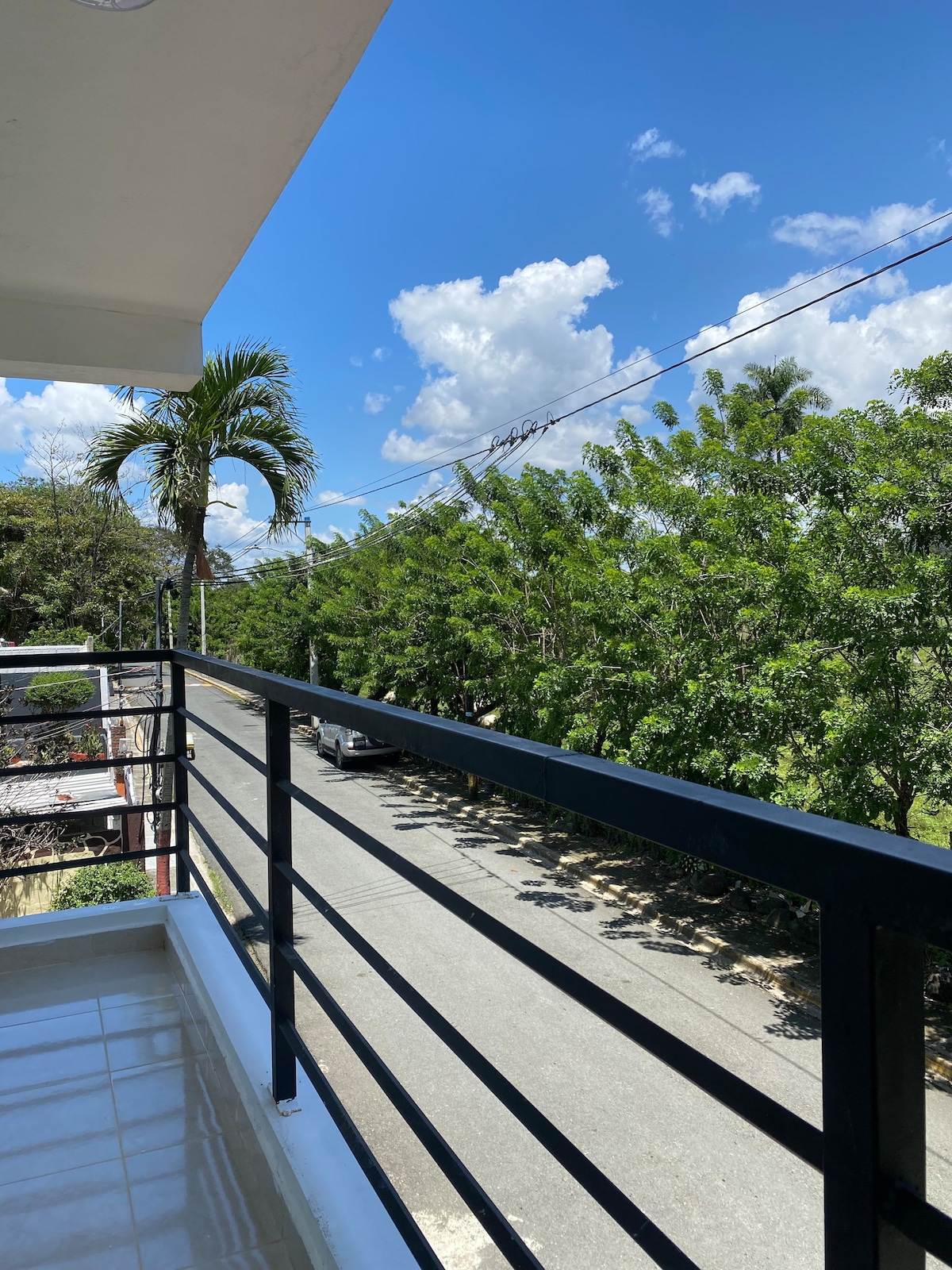 A balcony view features a railing overlooking a tree-lined street under a blue sky filled with fluffy clouds. Lush greenery adds to the tranquil atmosphere, while a glimpse of vehicles on the street can be seen in the distance.