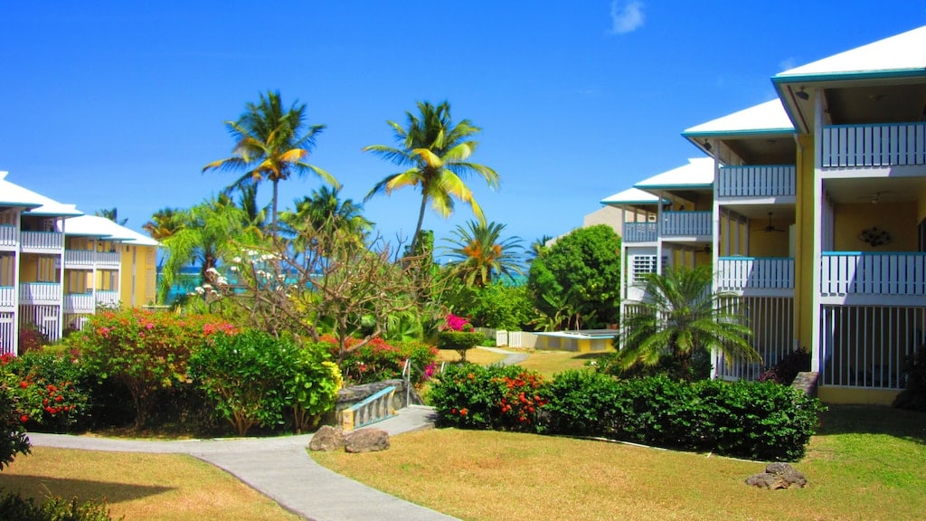 The view features a landscaped garden area surrounded by colorful tropical plants and palm trees. Two-story condominium buildings can be seen in the background, with balconies showcasing light-colored railings. A pathway winds through the garden, leading towards the ocean visible in the distance.