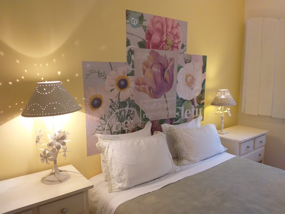 The cozy sleeping area features a queen-sized bed with light, neutral bedding. A floral mural serves as a backdrop. Two decorative lamps provide soft lighting on either side of the bed, complemented by light yellow walls that enhance the room's welcoming ambiance.