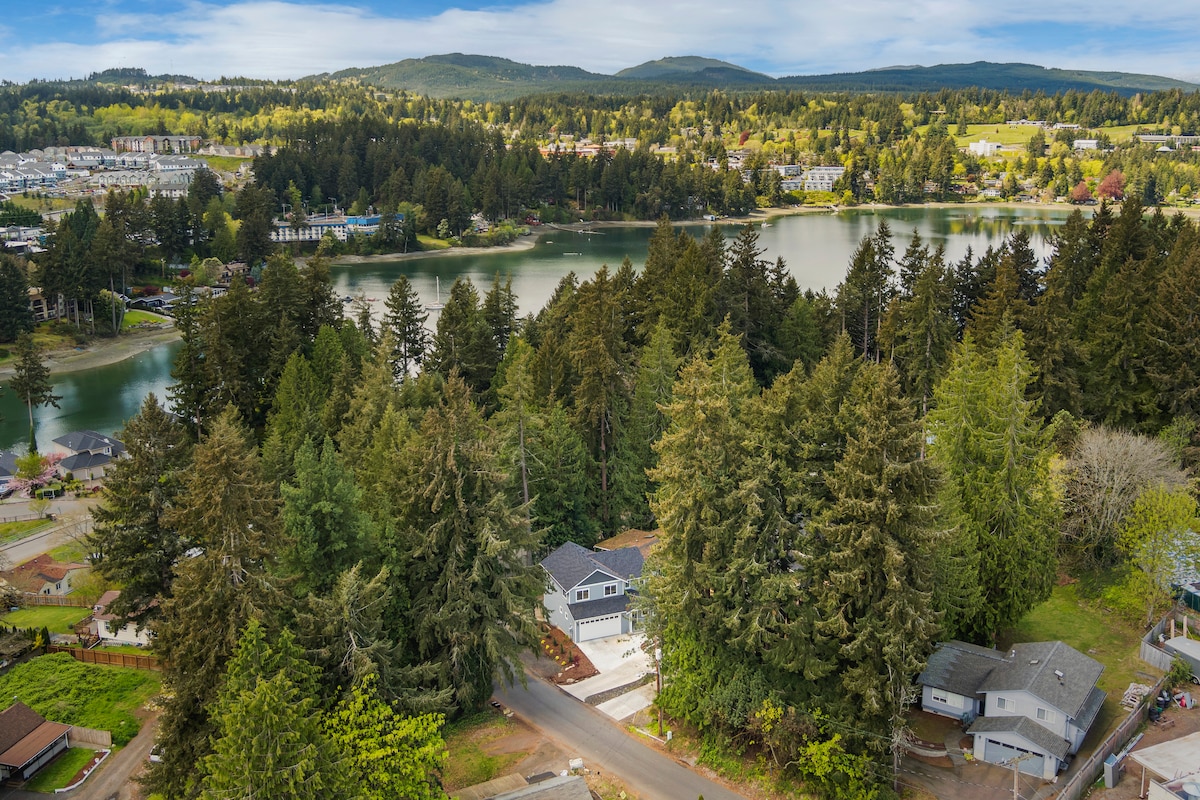 Aerial view captures a new home nestled among tall trees with a serene lake visible nearby. Expansive green landscapes and a variety of trees create a natural backdrop, showcasing the peaceful surroundings of the property.