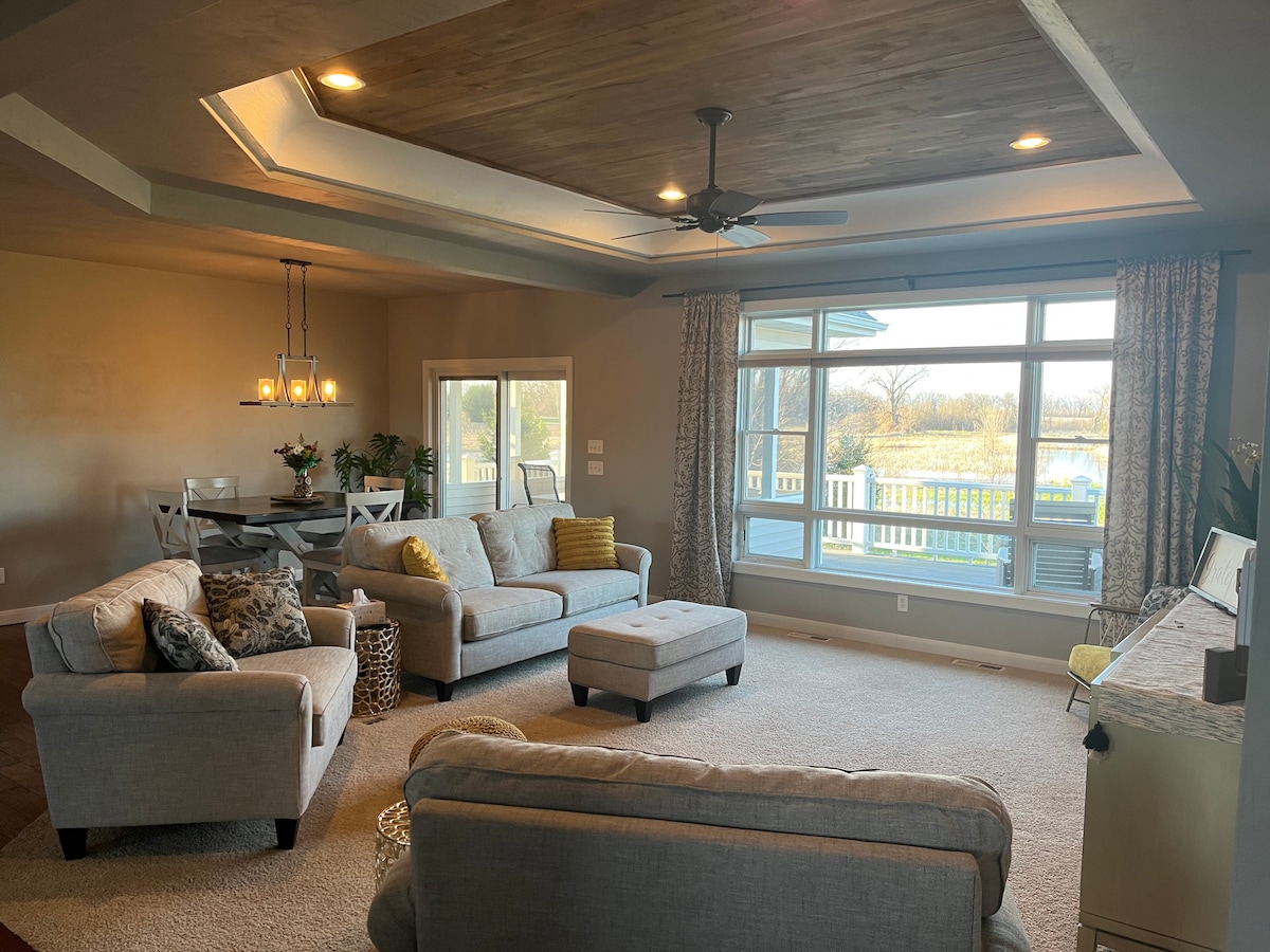 A spacious living area showcases a comfortable gray sectional and an ottoman. Large windows provide a view of the serene outdoor landscape. A dining table set for four is positioned nearby, illuminated by pendant lighting. Warm wood accents are featured in the ceiling design.