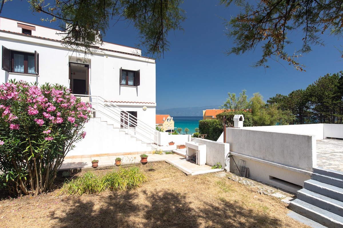 A white building with dark window frames is set against a clear blue sky, surrounded by greenery. A garden area features flowering bushes and a neat pathway leading to the entrance. The sea can be glimpsed in the distance, adding to the serene atmosphere.