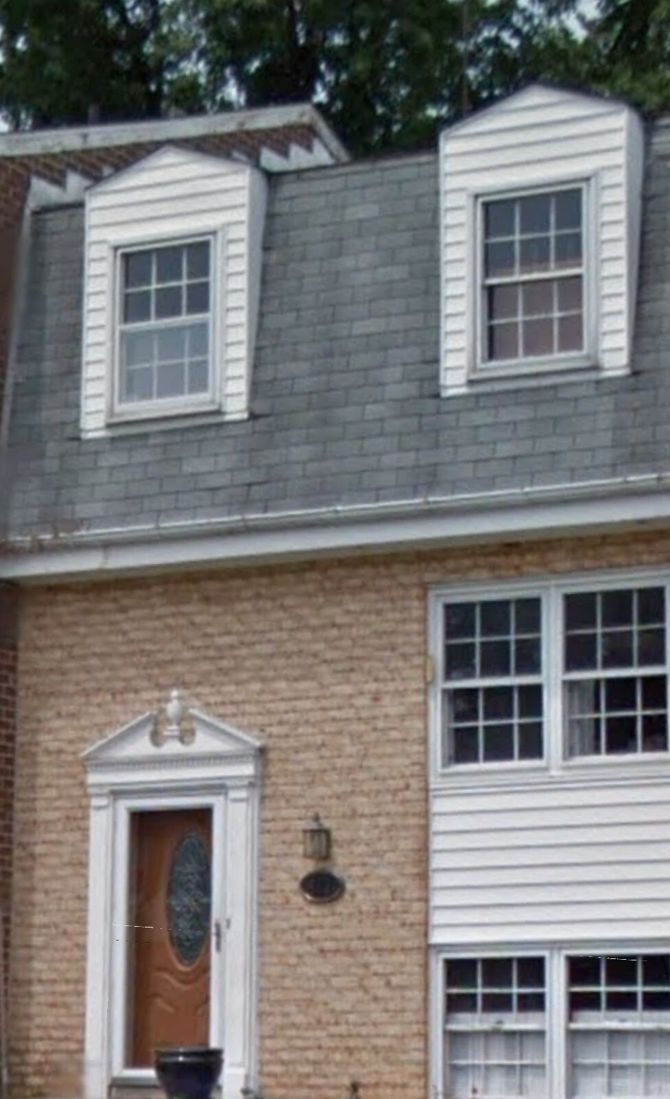 The exterior of a townhome is shown, featuring a combination of brick and siding. Two dormer windows are positioned above the main entrance, which showcases a decorative door with an oval glass insert. A wall-mounted light fixture is visible beside the door.