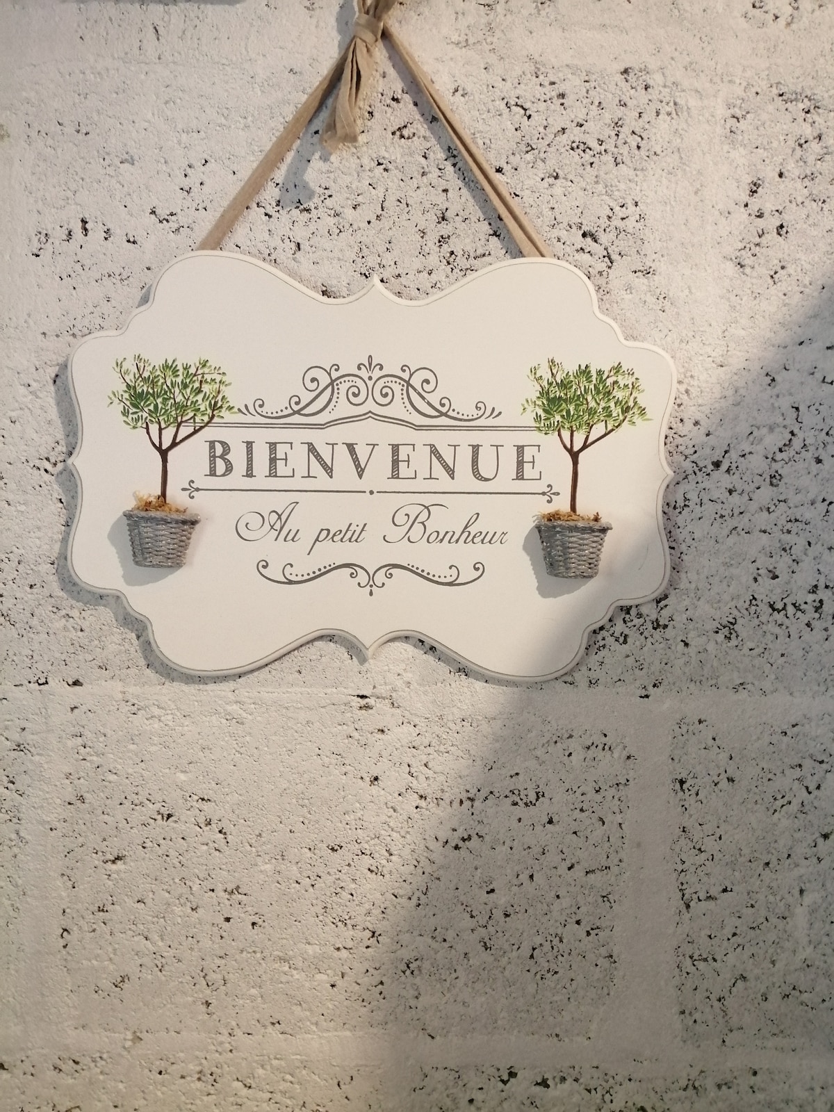 A welcome sign is displayed against a textured wall. The sign features decorative elements including two potted trees and the text 'Bienvenue Au petit Bonheur' in elegant font, adding a touch of charm to the entryway.