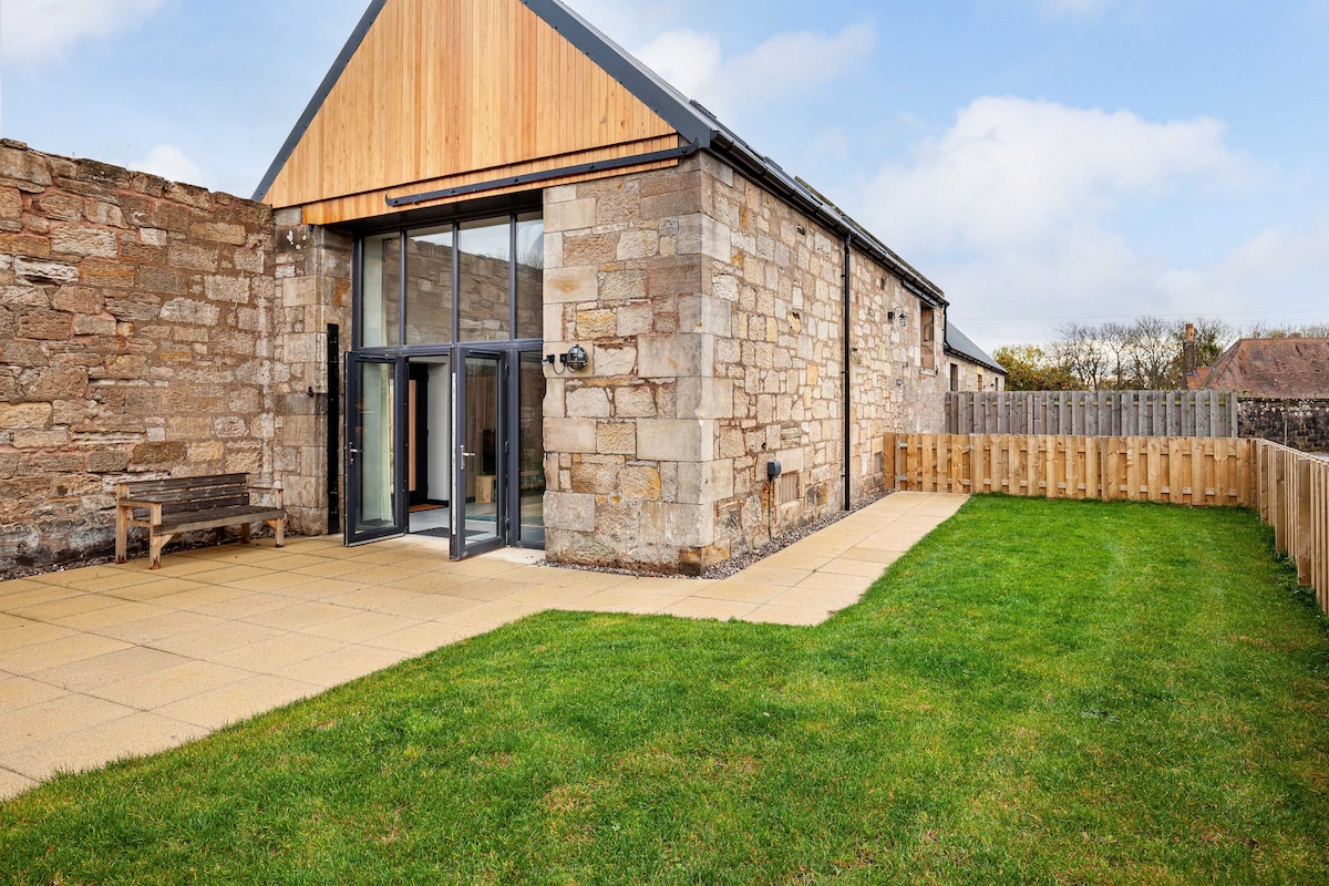 The exterior of a newly converted barn is featured, showcasing a mix of natural stone and wood. A spacious patio area with paved stones leads to a fully contained grassy yard, bordered by wooden fencing, creating a peaceful outdoor retreat.