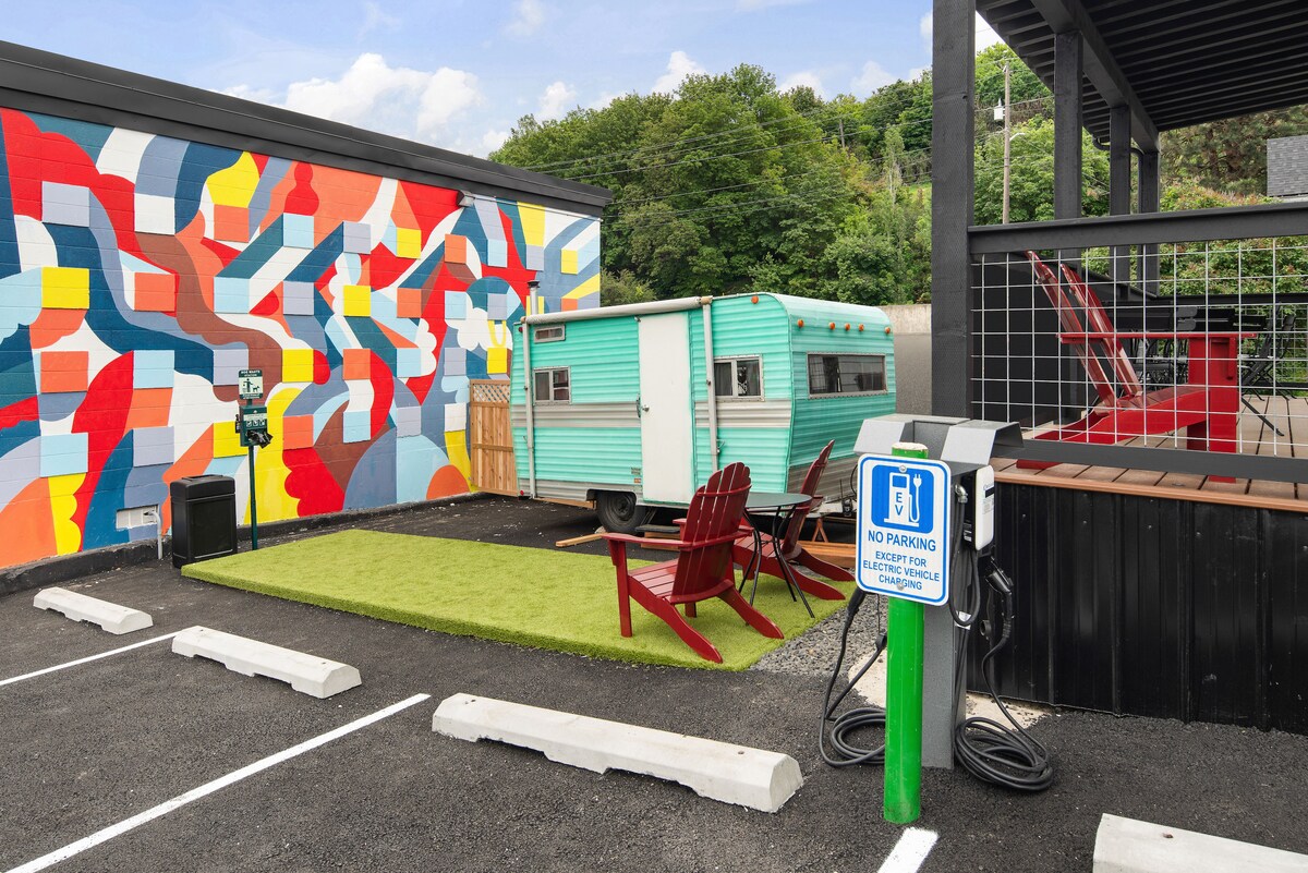 A vibrant mural adorns the exterior wall of a building, displaying an array of colors. In the foreground, two red Adirondack chairs are positioned on a patch of green grass beside a vintage teal trailer, creating a casual outdoor seating area.