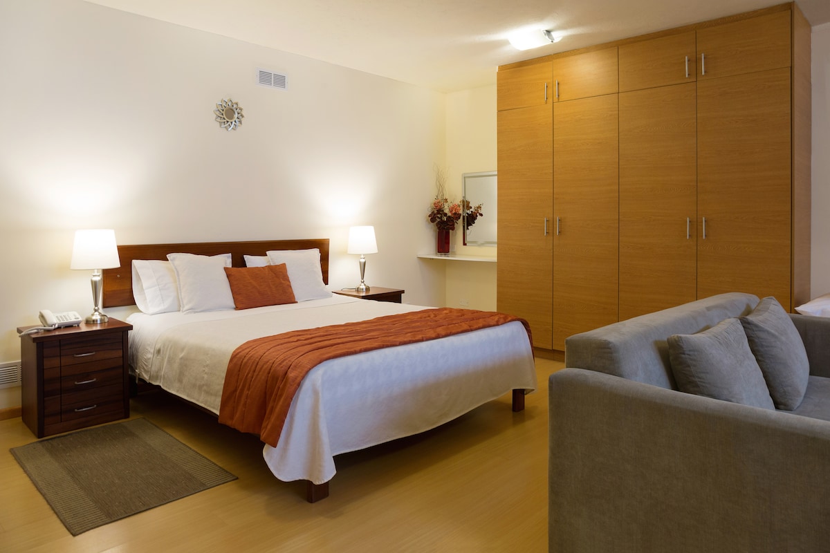 A spacious room features a queen bed with an orange accent pillow, complemented by two bedside tables with lamps. A gray sofa is positioned nearby. A large wooden wardrobe offers ample storage, while a wall-mounted mirror and decorative flowers add a touch of elegance.