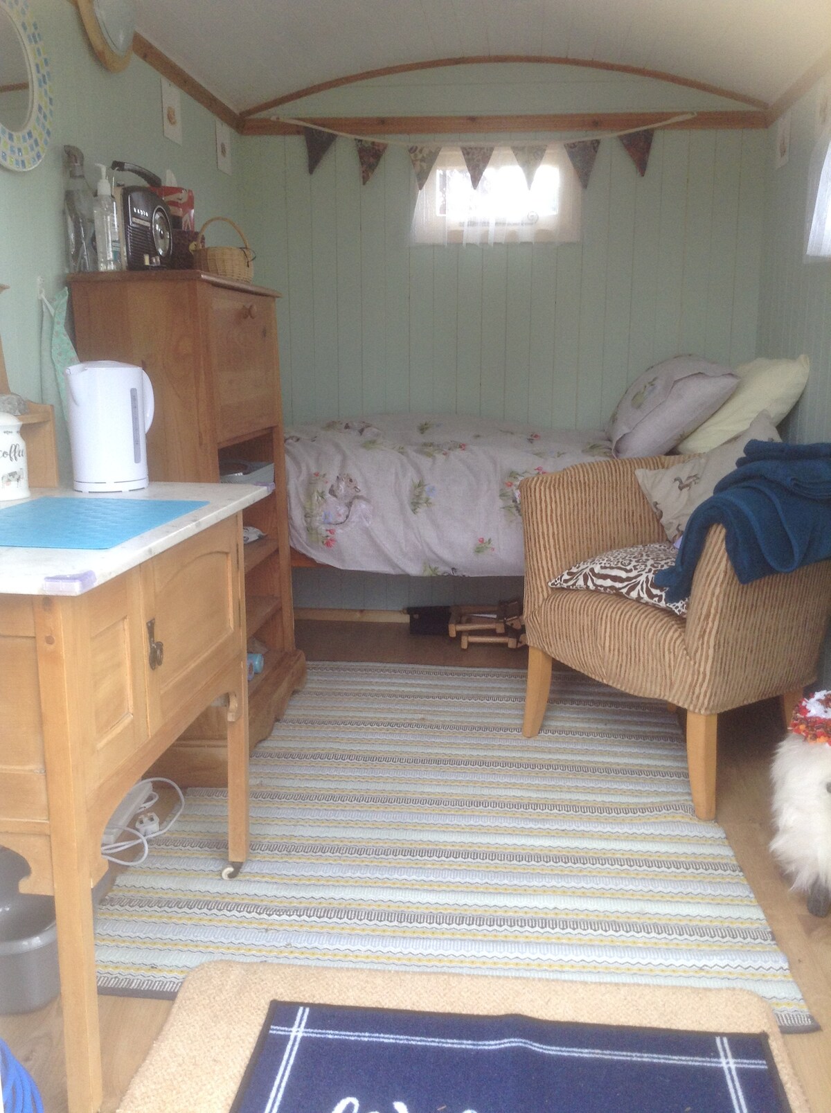 The interior of a cozy retreat features soft green walls and natural wood accents. A spacious bed is adorned with floral bedding, while a comfortable armchair offers a relaxing seating option. A small kitchenette with a kettle and wooden cabinet is visible, enhancing functionality.