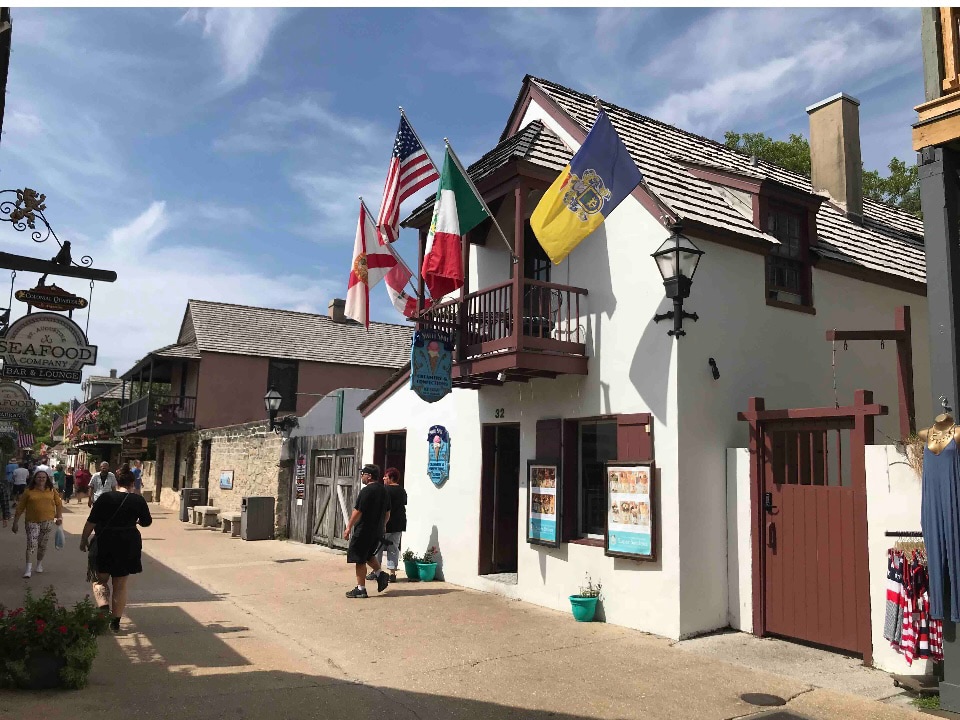 The image features a historic building in St. Augustine, adorned with multiple flags, including those of the United States, Italy, and Florida. The entrance is framed by a decorative lamp post, and pedestrians are seen walking along the pedestrian-only street, contributing to the lively atmosphere.