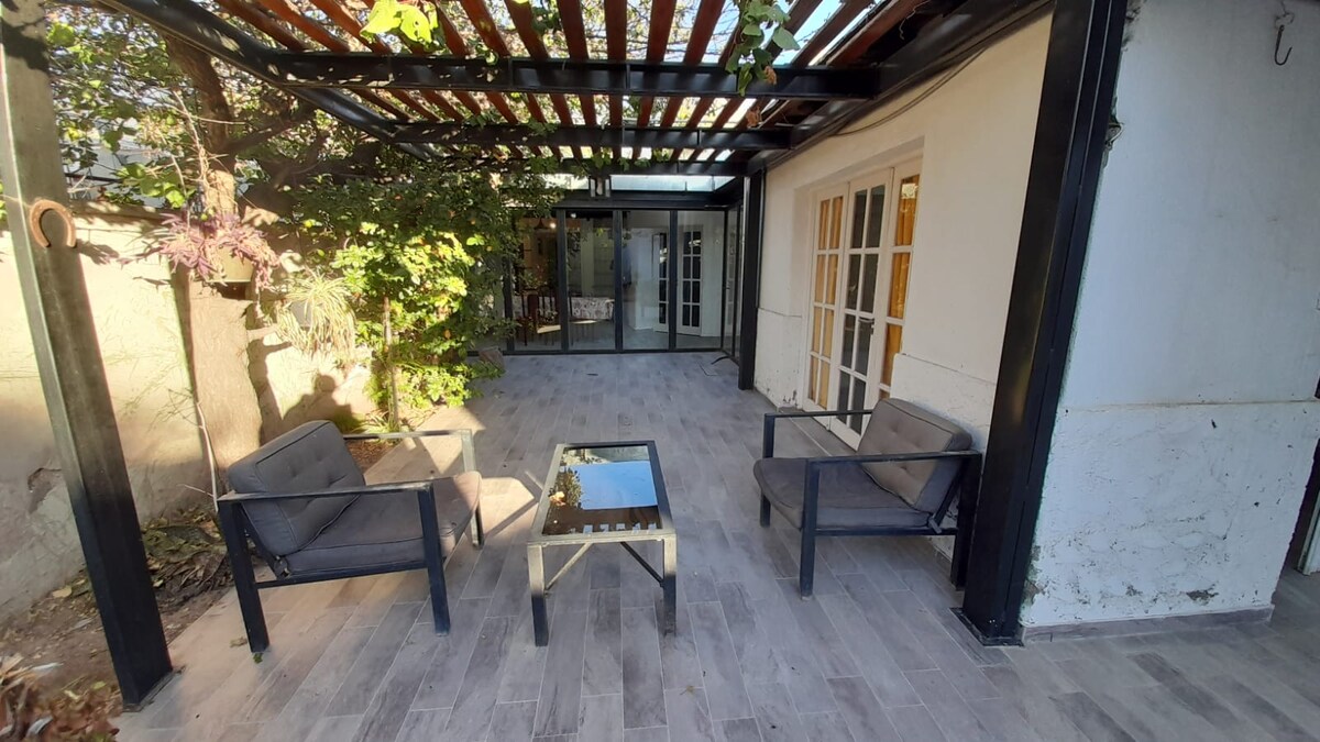 An outdoor seating area features two cushioned chairs and a glass-topped table, surrounded by greenery. The space is partially covered, allowing for natural light to filter through while offering a sheltered environment. Nearby, large glass doors lead into the interior of the house.