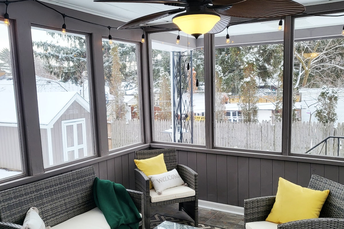 A sunroom is displayed with large windows offering views of the snowy outdoors. Two cushioned chairs in woven material are positioned with a decorative yellow pillow on one. A green throw blanket is draped casually over the arm of one chair, and a ceiling fan is visible above.