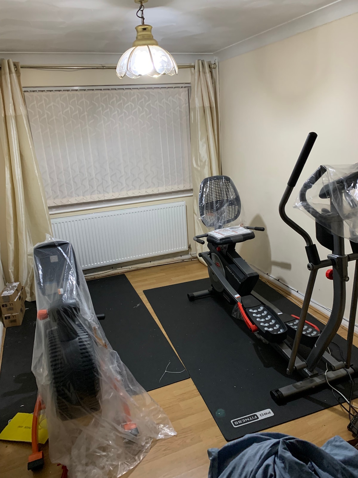 A well-equipped gym area is presented, featuring two exercise machines covered with protective plastic. The space includes wooden flooring and is illuminated by a pendant light. A large window with sheer curtains allows natural light to enter, enhancing the workout environment.
