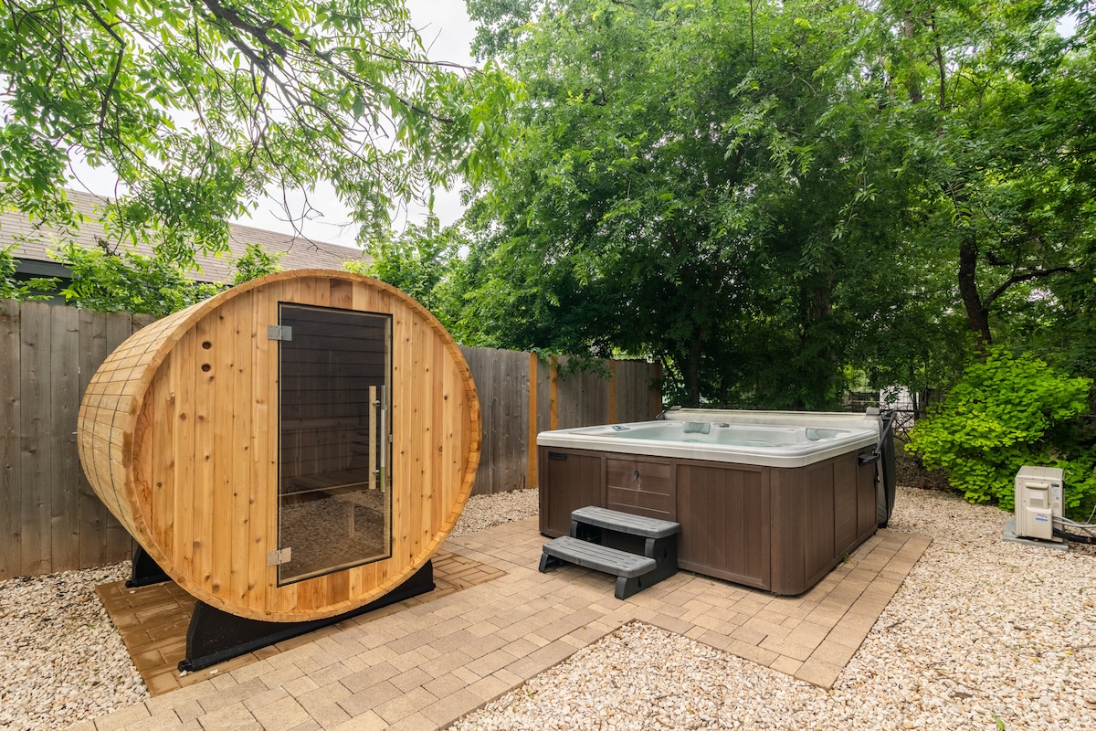 A wooden sauna cabin is positioned next to a hot tub, both nestled within a landscaped outdoor area. The sauna features a rounded design with a solid wood exterior, while the hot tub is rectangular and surrounded by light-colored stones. Lush greenery provides a natural backdrop.