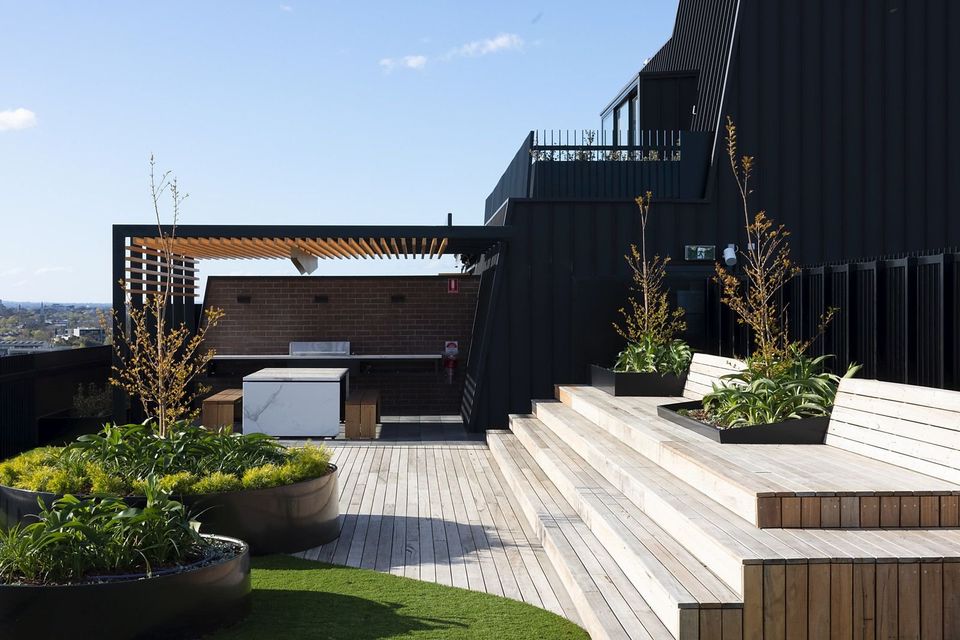 The communal rooftop area features wooden decking leading to a landscaped garden with planters and steps. Outdoor seating arrangements are visible, accompanied by a BBQ area under a shaded pergola. Plants and greenery enhance the space, offering an inviting setting for relaxation.