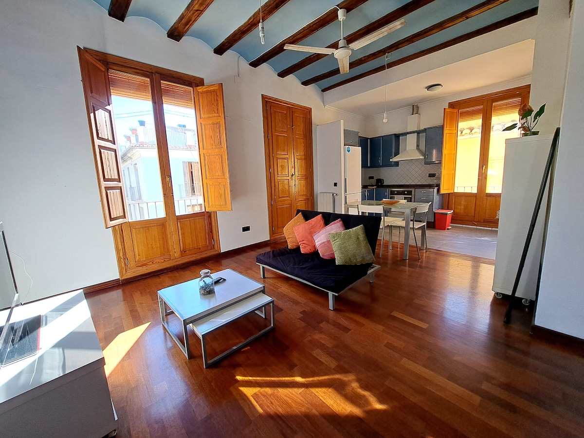 The airy living area features tall wooden windows allowing natural light to fill the space. A dark seating area with colorful cushions is complemented by a simple coffee table. An integrated kitchen area is visible, connecting to a dining table. Light wooden beams are present in the ceiling.