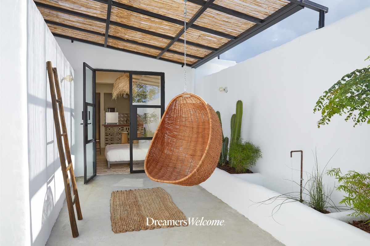 A cozy outdoor space features a welcoming hanging chair made of natural fibers, complemented by a soft rug. Lush greenery, including cacti, adds a touch of nature. The open doorway leads to a bedroom, with warm light filtering in through tall windows.