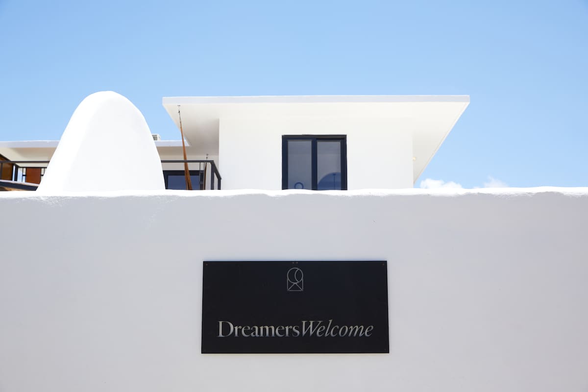 A clean white wall surrounds the property, featuring a modern sign that reads 'DreamersWelcome.' The bright sky above creates a serene backdrop for the building, which is characterized by minimalist architecture and large windows.