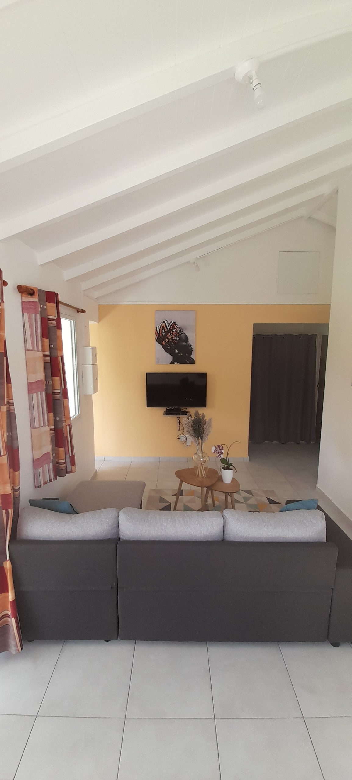 A spacious living area is highlighted by a comfortable gray sofa, accompanied by decorative cushions. A coffee table sits at the center, while an art piece adorns the wall above. A flat-screen television is mounted, and natural light fills the room through large windows.