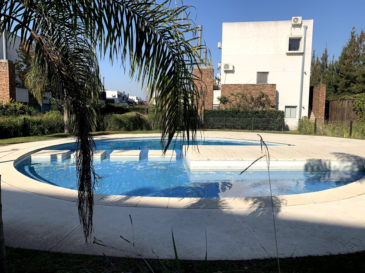 A swimming pool is set within a landscaped area, surrounded by greenery. The circular pool features a shallow section and is framed by a smooth concrete deck. Residential buildings are visible in the background, providing a sense of privacy and tranquility.