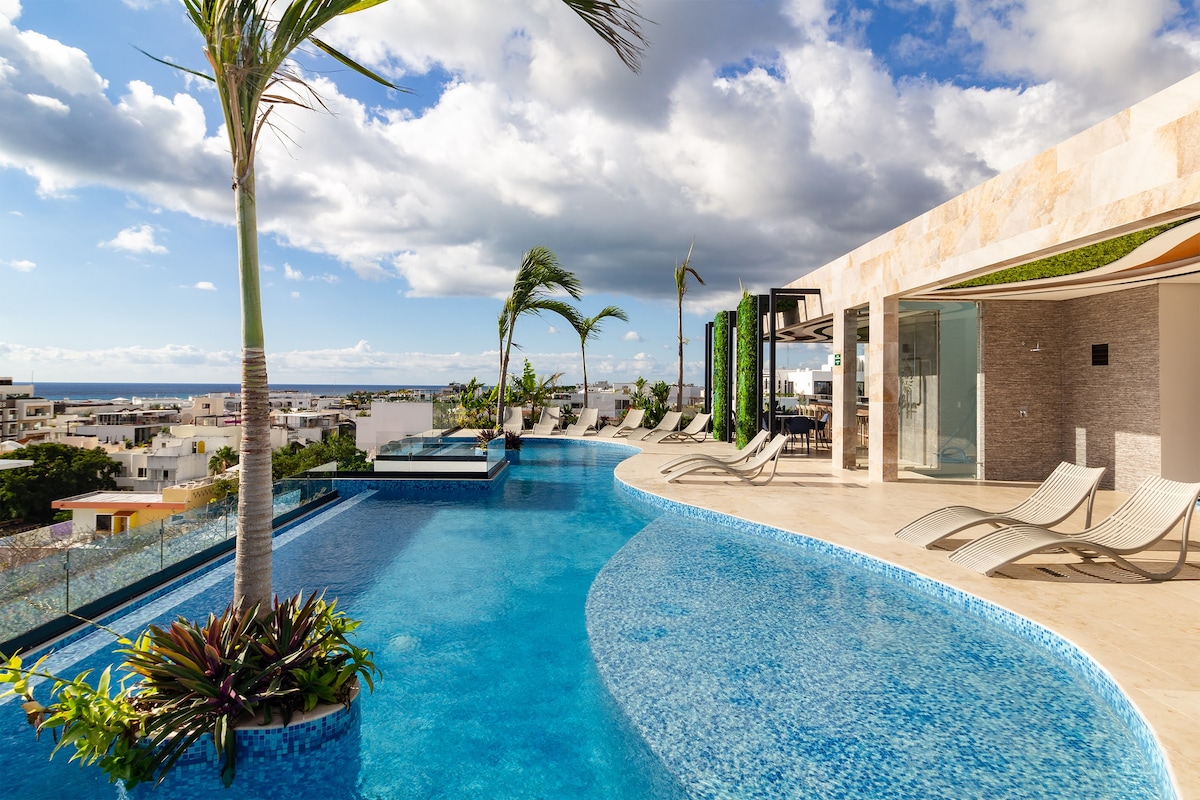 A rooftop pool area offers expansive views of the surrounding city and ocean. The shimmering blue water of the pool reflects the sky, while sun loungers are arranged along the edge. Lush greenery lines the pool, enhancing the serene atmosphere.