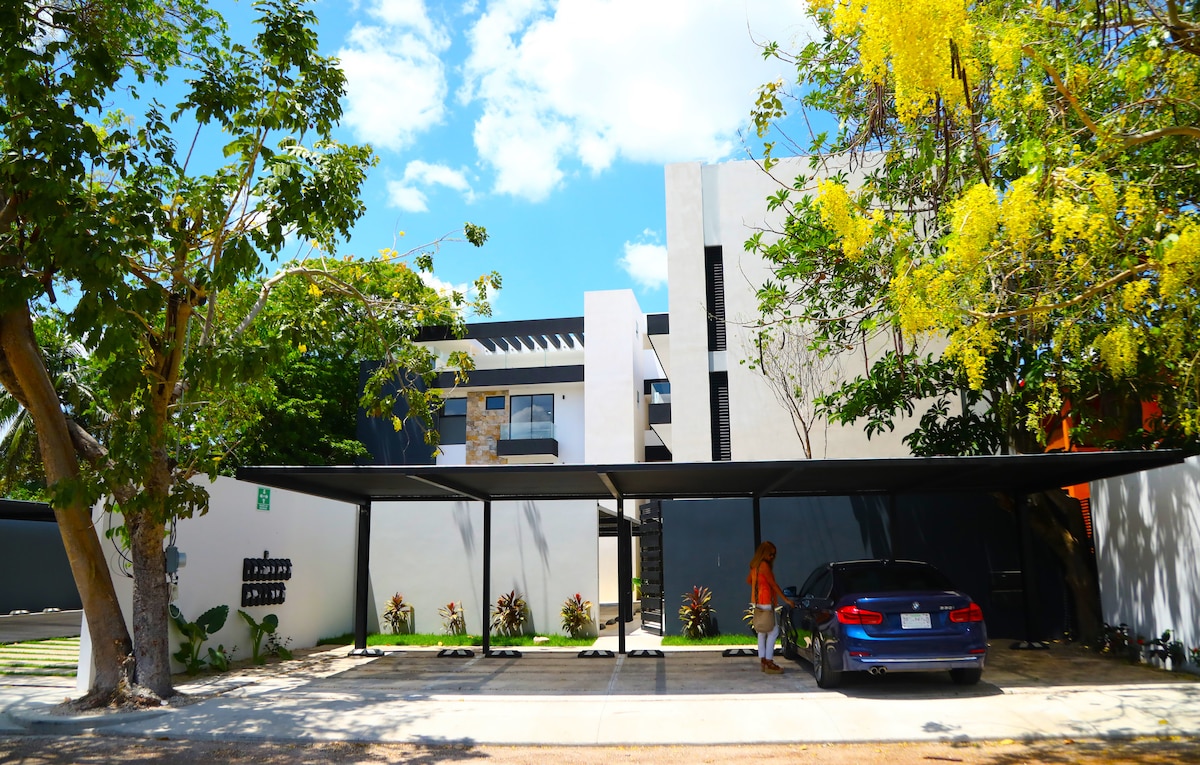 An exterior view of the condominium features a modern design with white and gray walls. A spacious carport shelters a parked vehicle, surrounded by well-maintained landscaping and several green trees. The bright blue sky enhances the contemporary feel of the building.