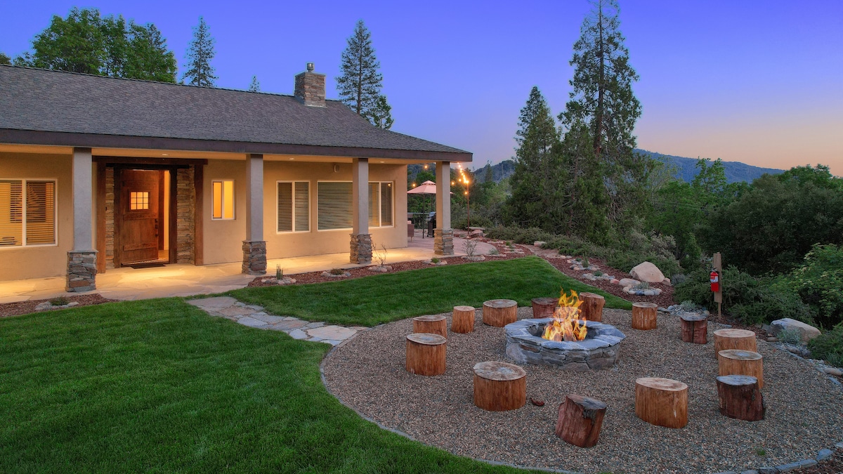 An inviting outdoor space features a stone fire pit surrounded by log seating. Neatly trimmed grass and a stone pathway lead to the front porch of the home, where warm light shines from inside. Trees and hills create a serene backdrop against the evening sky.