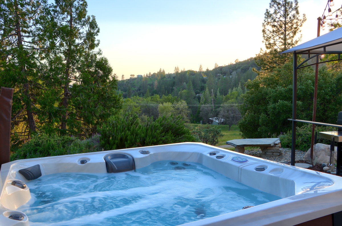 A well-maintained hot tub is positioned on a patio, offering a view of the surrounding greenery and distant hills. The bubbling water is visible, inviting relaxation amidst the tranquil outdoor setting. The area is framed by trees and natural landscaping, enhancing the serene atmosphere.