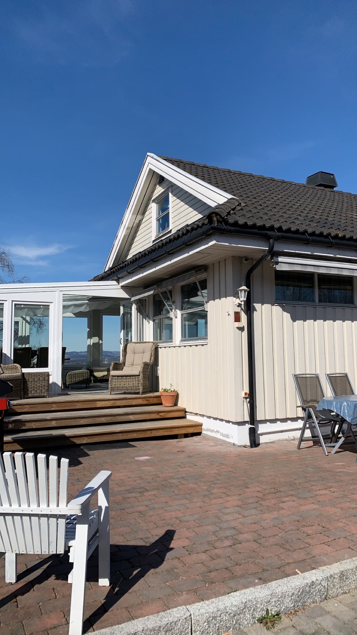 The exterior of the house features a light-colored facade and a steep roof. A spacious wooden deck leads to a glass-enclosed garden room, while outdoor seating options, including a few patio chairs, provide inviting spaces to relax. The sky above is clear and bright.