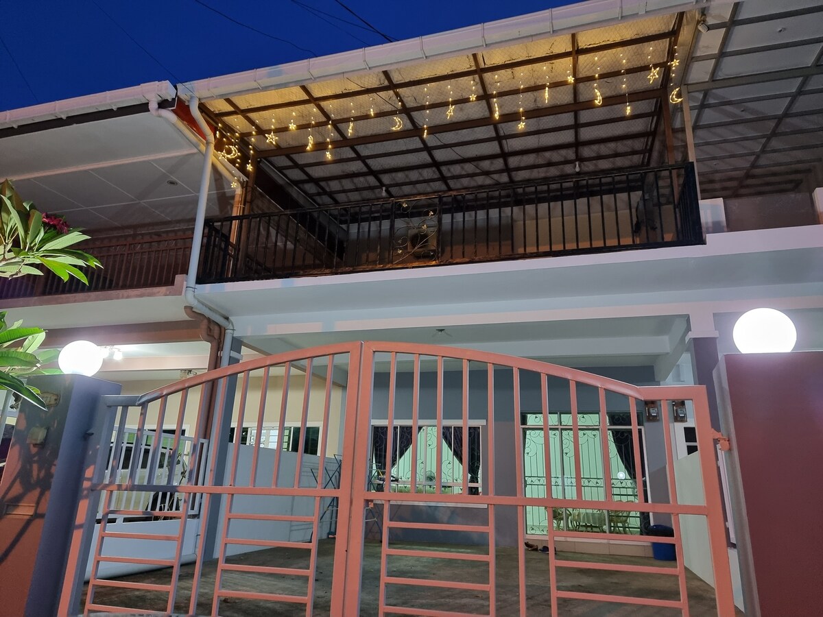 A two-story building is presented, featuring a covered balcony adorned with string lights. A gate welcomes guests, with soft landscaping visible at the front. The ground level shows large windows, allowing natural light to illuminate the interior spaces.