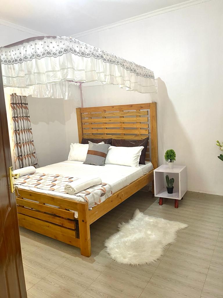 The bedroom features a wooden bed with an elegant canopy above. Soft linens and decorative pillows enhance the bed's inviting look. A small side table stands beside the bed, complemented by a decorative plant. A plush rug adds warmth to the wooden floor.