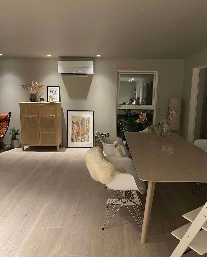 A modern dining area is showcased, featuring a long wooden table surrounded by white chairs. An air conditioning unit is mounted on the wall, and a decorative cabinet holds various items. A large piece of artwork is displayed, enhancing the inviting environment.
