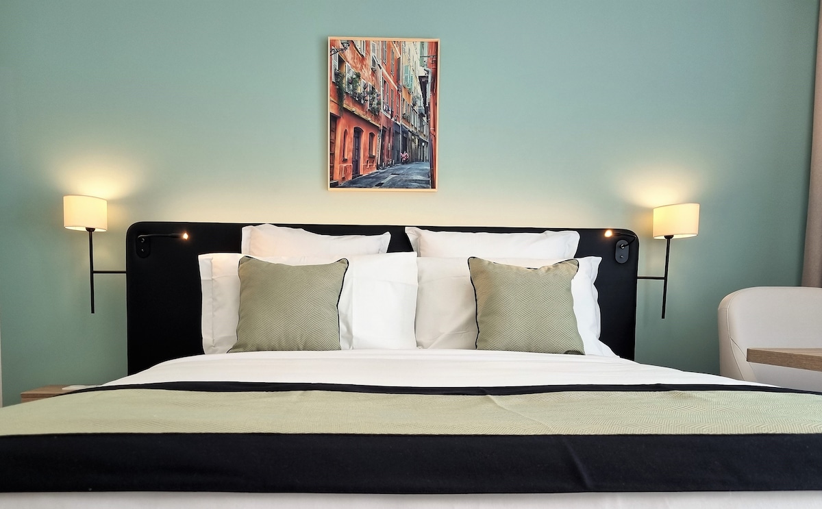 A spacious bed with white linens and two green-accented pillows is centered against a light green wall. A piece of artwork depicting a street scene hangs above the bed, flanked by two stylish bedside lamps that provide soft lighting.