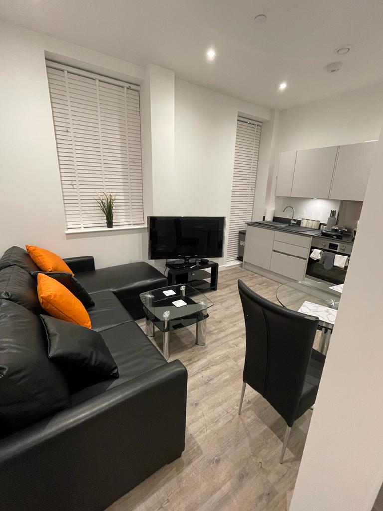 A modern living area features a black leather sofa with orange accent pillows. A glass table is set with two chairs, and a television is positioned against the wall. The space is complemented by large windows with vertical blinds, allowing natural light to enhance the ambiance.
