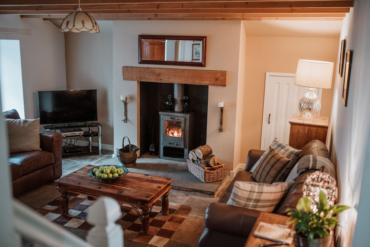 The living room features a log-burning stove set within a traditional stone fireplace, providing warmth. Comfortable seating options surround a rustic coffee table, while a TV is positioned nearby. Natural light fills the space through a window, enhancing the cozy atmosphere.