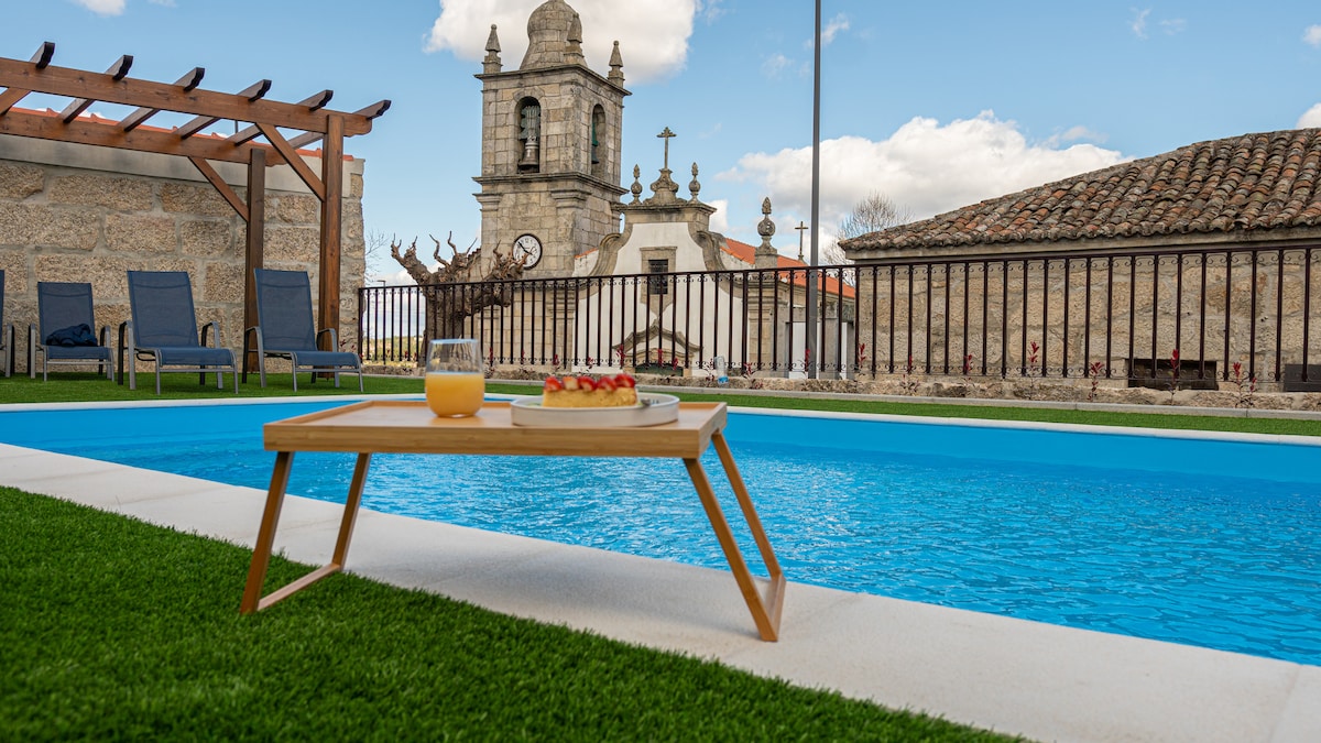 An outdoor area features a refreshing pool surrounded by well-maintained grass. A wooden table displays a drink and snacks, while lounge chairs are arranged nearby. In the background, a historic building and a clock tower add character to the setting.