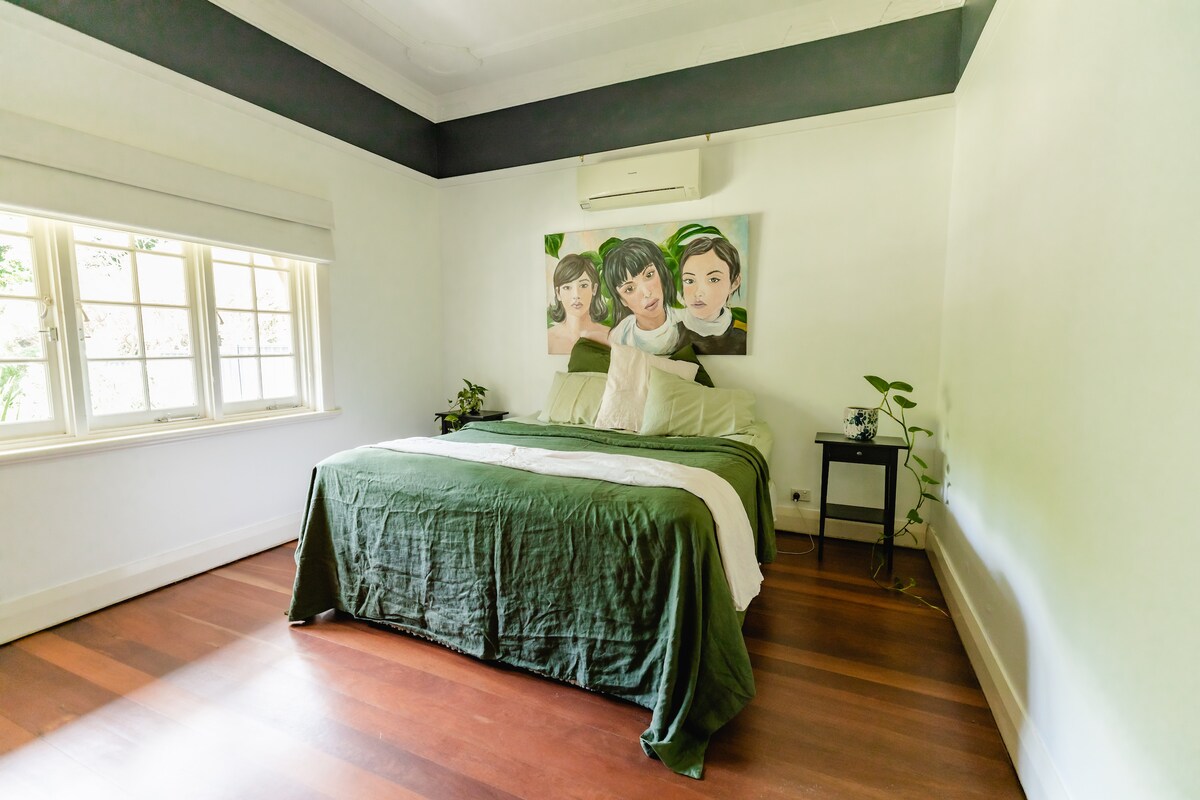 A spacious bedroom features a large bed with an olive green coverlet, complemented by cream-colored pillows. A small bedside table holds a lamp, and a potted plant adds a touch of greenery. Natural light filters through a window with soft shades.